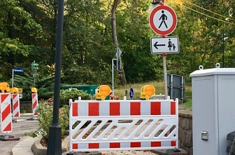 Baustelle_BF Auf dem Bild ist eine Baustelle im Außenbereich zu sehen. Mehrere rot-weiße Absperrungen blockieren einen Gehweg oder eine Straße. Auf den Absperrelementen befinden sich gelbe Warnleuchten. Rechts steht ein Verkehrsschild, das Fußgängern den Durchgang verbietet; darunter zeigt ein Zusatzschild mit Pfeil nach links, dass der Fußweg auf die andere Seite umgeleitet wird. Im Hintergrund sind viele grüne Bäume, ein paar Sträucher und einzelne Gebäude zu erkennen. Die Szene wirkt wie ein ruhiger Bereich am Rand eines Wohn- oder Parkgebiets.