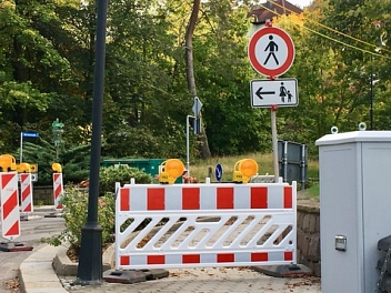 Auf dem Bild ist eine Baustelle im Außenbereich zu sehen. Mehrere rot-weiße Absperrungen blockieren einen Gehweg oder eine Straße. Auf den Absperrelementen befinden sich gelbe Warnleuchten. Rechts steht ein Verkehrsschild, das Fußgängern den Durchgang verbietet; darunter zeigt ein Zusatzschild mit Pfeil nach links, dass der Fußweg auf die andere Seite umgeleitet wird. Im Hintergrund sind viele grüne Bäume, ein paar Sträucher und einzelne Gebäude zu erkennen. Die Szene wirkt wie ein ruhiger Bereich am Rand eines Wohn- oder Parkgebiets.&nbsp;&copy;&nbsp;Anja Seidel