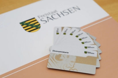 Ehrenamtskarte_BF Auf dem Bild liegt eine Mappe mit dem Wappen des Freistaates Sachsen und der Aufschrift „Freistaat Sachsen“. Darauf sind mehrere nebeneinander ausgebreitete Karten zu sehen. Die Karten tragen den Schriftzug „Ehrenamtskarte“ und zeigen ebenfalls das sächsische Wappen. Die Farben im Bild sind überwiegend Weiß, Beige und leichte Gold. © Sächsisches Staatsministerium für Soziales