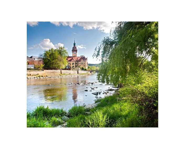 Das Bild zeigt das Rathaus von Waldheim an der Zschopau.
Das Rathaus ist ein großes, historisches Gebäude mit gelber Fassade und rotem Dach. In der Mitte steht ein hoher Turm mit einer Uhr und einer dunklen Turmspitze. Das Gebäude liegt direkt am Ufer.
Im Vordergrund sieht man den Fluss. Das Wasser ist ruhig, und einige Steine ragen aus dem Wasser heraus. Am Ufer wächst grünes Gras. Rechts im Bild hängt ein großer Baum mit langen, grünen Zweigen über das Wasser.
Der Himmel ist blau mit weißen Wolken. Das Sonnenlicht lässt das Rathaus und die Natur hell und freundlich wirken.
Das Bild zeigt eine ruhige, idyllische Landschaft mit dem Rathaus von Waldheim im Mittelpunkt.